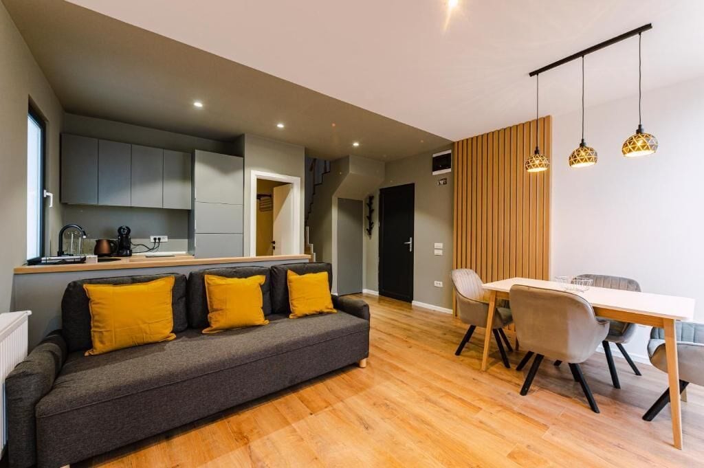 A modern living room with a grey sofa, yellow cushions, a wooden dining table with chairs, and pendant lighting.