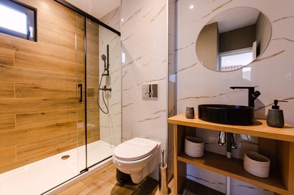 Un baño moderno con ducha de vidrio, inodoro, tocador con lavamanos negro y espejo redondo.