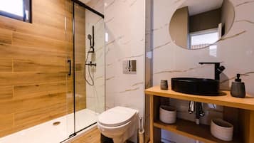 Un baño moderno con ducha de vidrio, inodoro, tocador con lavamanos negro y espejo redondo.