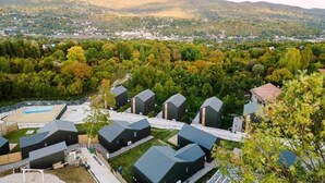 Zona residencial amb cases modernes de color fosc i teulades inclinades, envoltada de vegetació exuberant i turons al fons.