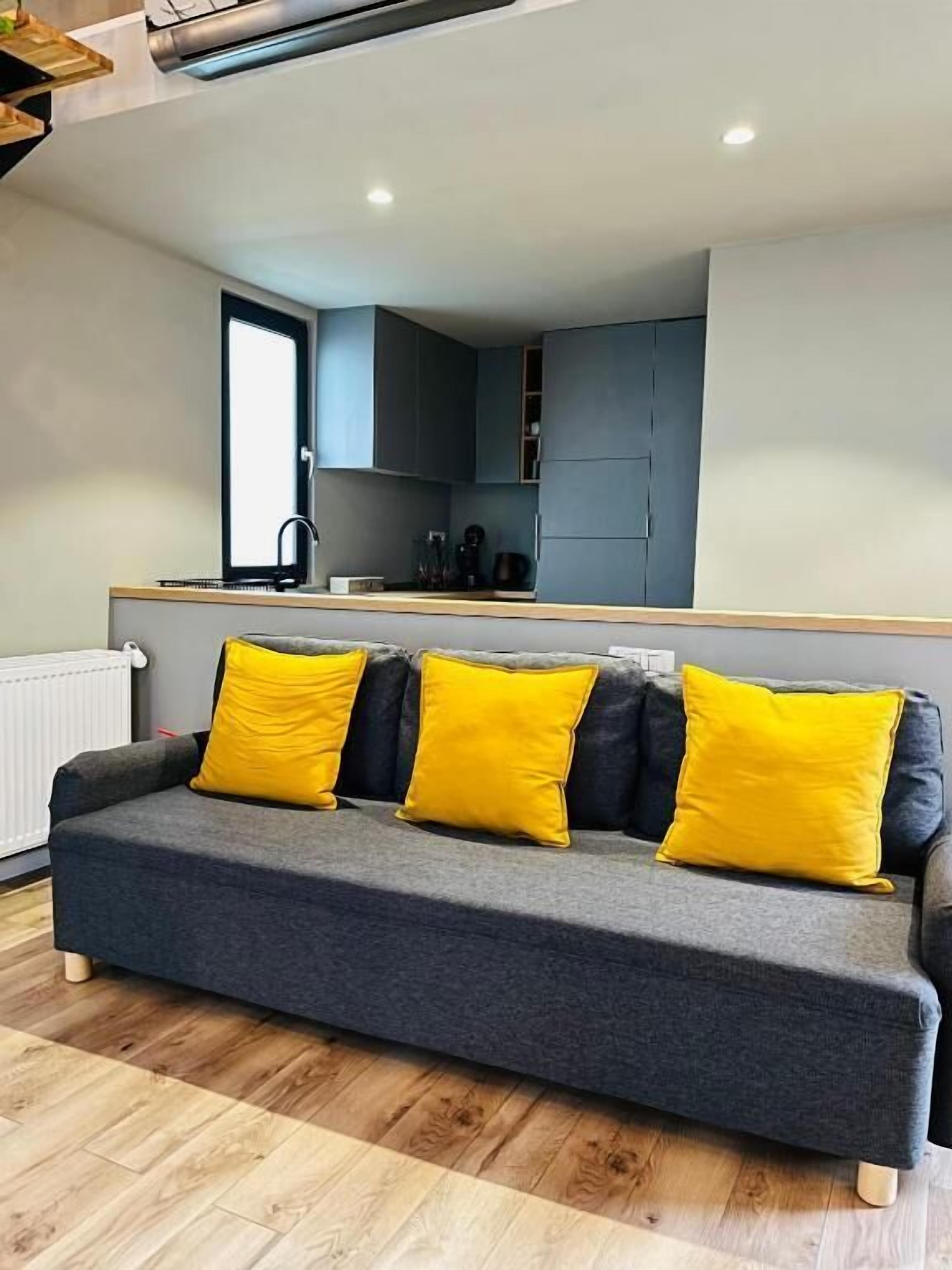 A modern living room with a grey sofa and yellow cushions, a wooden floor, and a kitchen area with grey cabinets and a window above the sink.