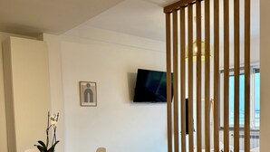 A modern hotel room with a wooden partition, a dining area with a table and chairs, a flat-screen TV mounted on the wall, and a bed with a view.