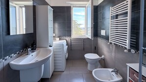 A modern bathroom with a bidet, toilet, and sink, a large mirror, and a view through a window.
