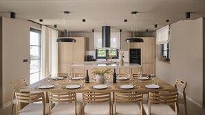 A modern dining area with a large wooden table set for six, surrounded by wooden chairs with cushioned seats. There are pendant lights above the table, and a kitchen area is visible in the background. - Villa Madora in Porec (Porec)