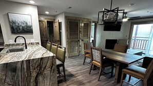 A modern kitchen with a marble island, wooden dining table, and chairs. There is a painting on the wall and a large window with a view.
