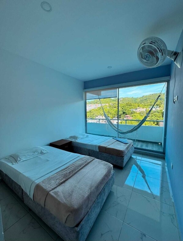 A bedroom with two beds, a hammock, a large window with a view, and a ceiling fan.