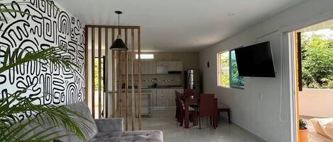 A modern living room with a sofa, a coffee table, and a TV. There is a kitchen in the background and a sliding glass door leading to an outdoor area.