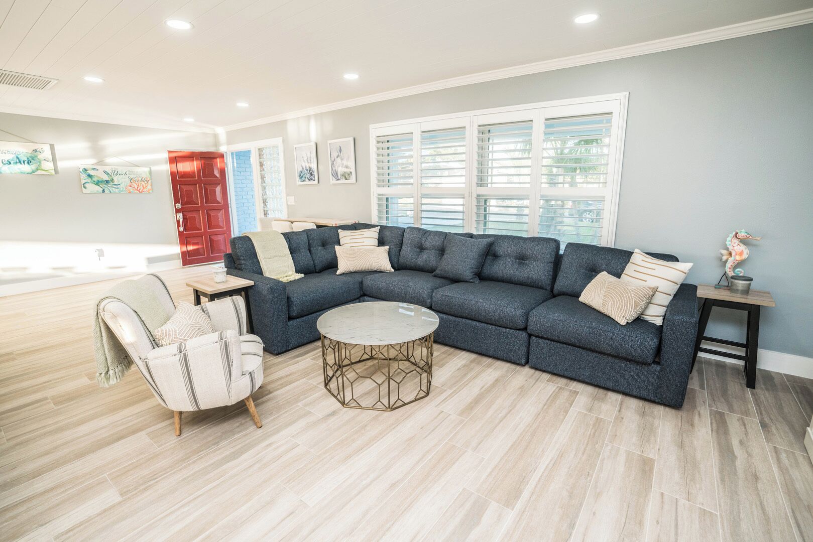 A modern living room with a sectional sofa, a round coffee table, and a chair. There is a red door and a window with blinds.