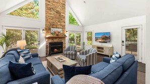 A spacious living room with a stone fireplace, large windows, and a television. - Juniper Presents: Oasis in the Woods - Hot Tub, Pool & Pizza Oven (New Buffalo)
