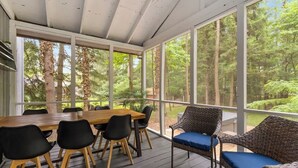 Un espacio al aire libre techado con una mesa y sillas de madera, ventanas amplias y vista a un bosque.