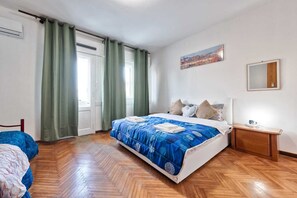 A bedroom with a large bed, wooden flooring, a nightstand, a mirror, and a window with curtains.