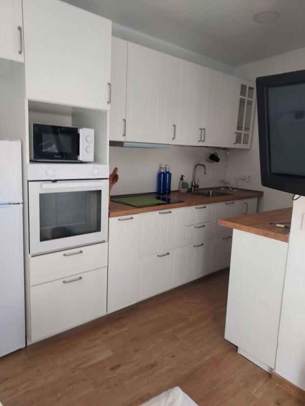 Una cocina compacta con armarios blancos, un microondas, un horno, un fregadero y un televisor.