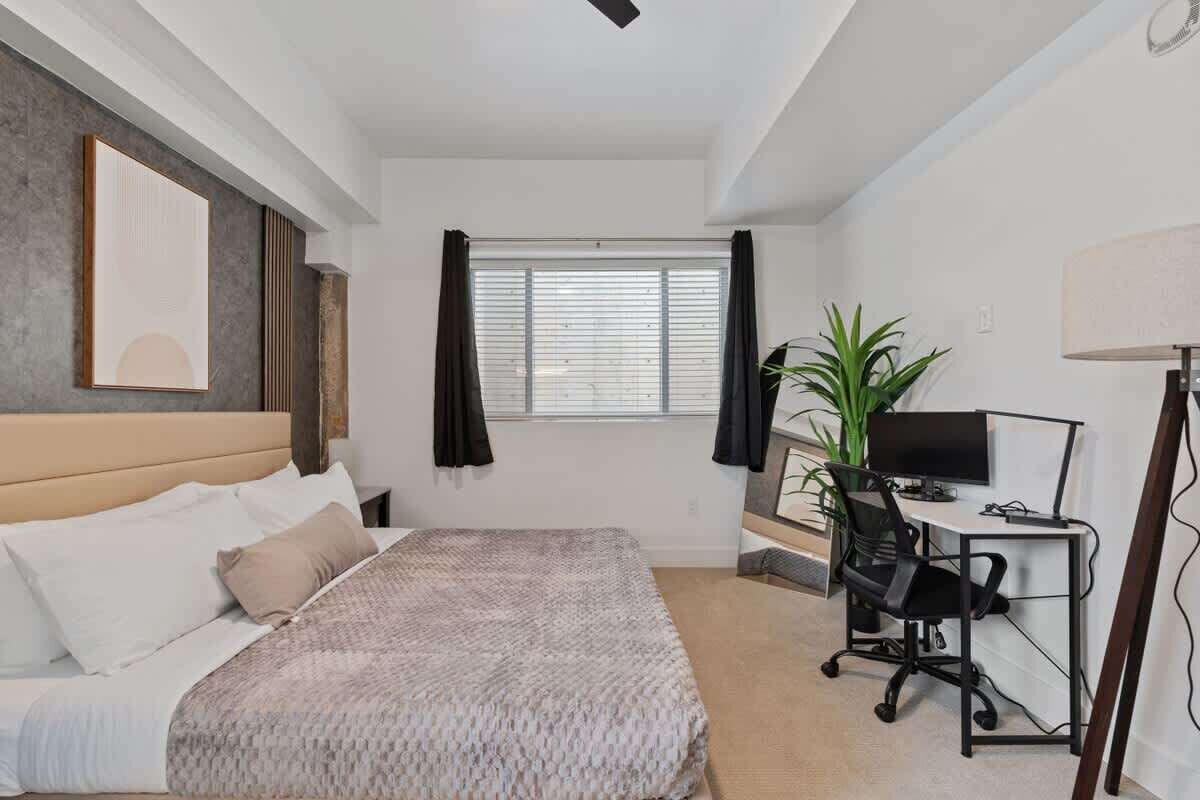 Une chambre à coucher avec un lit, un bureau avec un ordinateur, une plante et une fenêtre avec des rideaux.