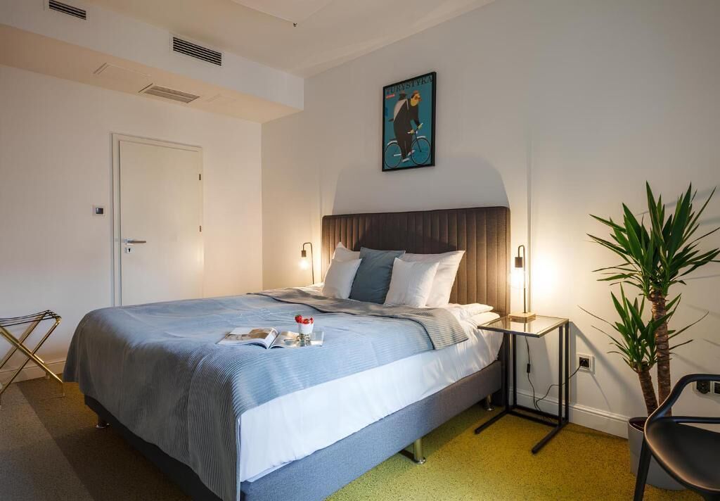 A neatly arranged hotel room with a large bed, bedside tables, a chair, a potted plant, and a door.