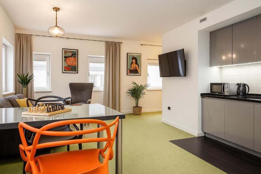 Una sala de estar moderna con una mesa de comedor de vidrio, sillas anaranjadas, un sofá, un televisor montado en la pared y una zona de cocina con armarios grises y una encimera negra.