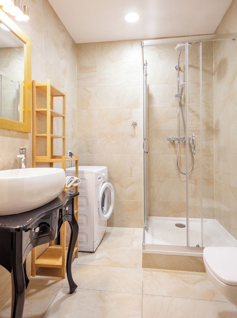 A modern bathroom with a sink, mirror, washing machine, and glass-enclosed shower.