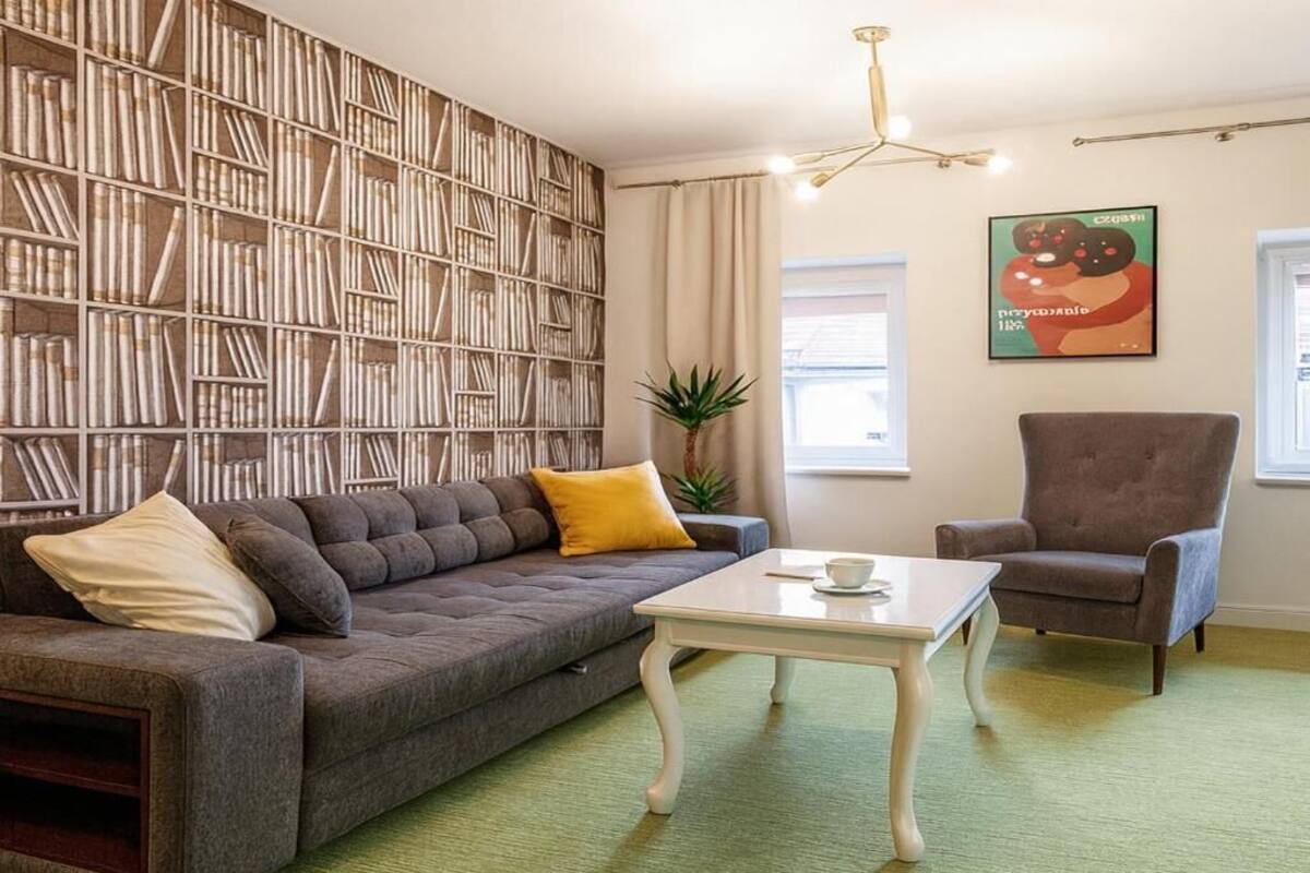 Ett vardagsrum med en grå soffa, ett vitt soffbord, en bokhylla, en brun fåtölj och en grön matta.