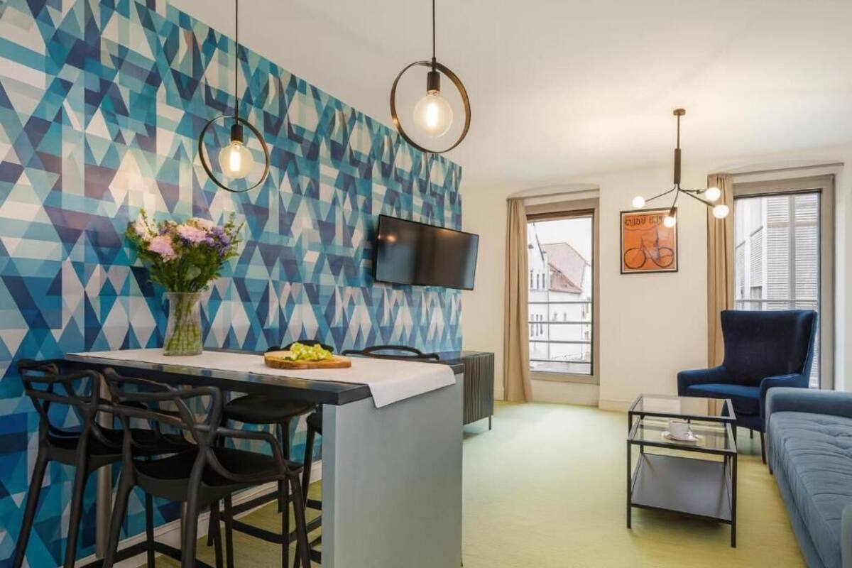 Ett modernt vardagsrum med en vägg dekorerad med ett geometriskt mönster, en platt-TV, en blå soffa, ett soffbord och en matplats med ett litet bord och stolar.