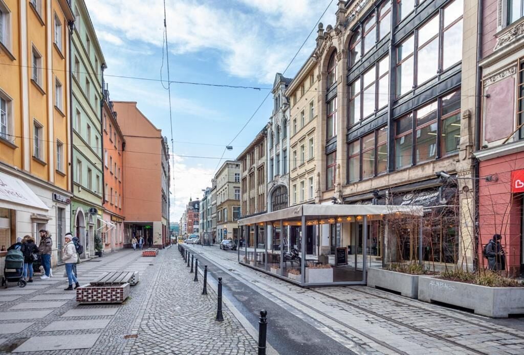 A cobblestone street lined with colorful buildings, a tram, and people walking.