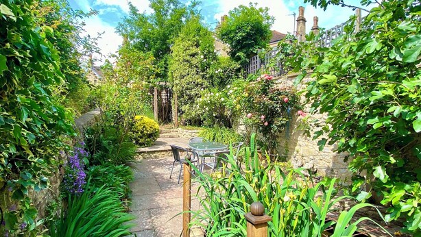 A garden pathway leading to a table and chairs, surrounded by lush greenery and a stone wall. - Charming 3 Bedroom House next to Victoria Park | Pass the Keys (Bath)