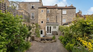 A stone building with a central door, a small porch, and a garden with various plants. - Charming 3 Bedroom House next to Victoria Park | Pass the Keys (Bath)