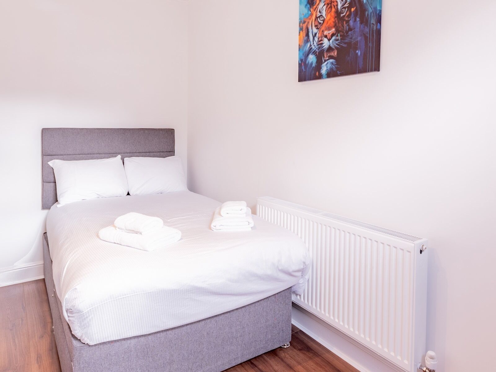 A neatly made bed with white linens and two folded towels, a grey upholstered headboard, and a white radiator.