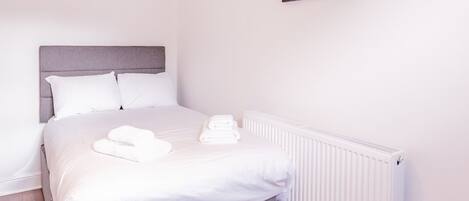 A neatly made bed with white linens and two folded towels, a grey upholstered headboard, and a white radiator.