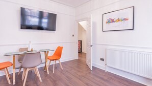A modern hotel room with a dining area featuring a glass table, wooden chairs, and a flat-screen TV mounted on the wall. - Pass the Keys | Comfortable Flat with Patio | Sleeps 4 (Westcliff-on-Sea)