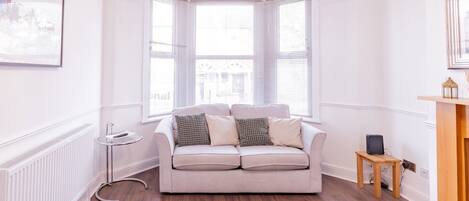Una habitación con un sofá color crema, dos ventanas con persianas, un radiador, una mesita auxiliar y un cuadro enmarcado en la pared.