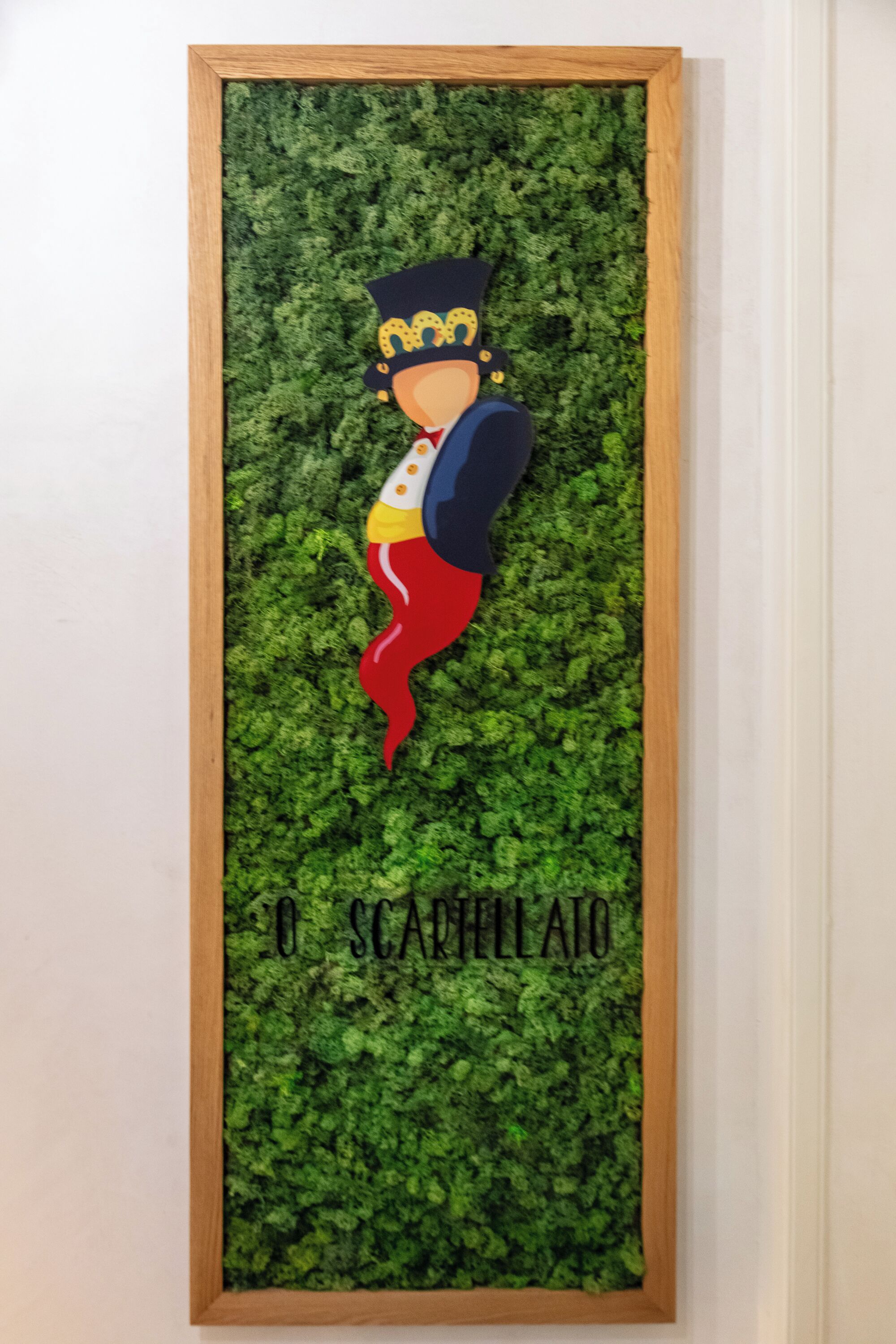 A framed artwork with a character wearing a crown and a red scarf, set against a green leafy background.