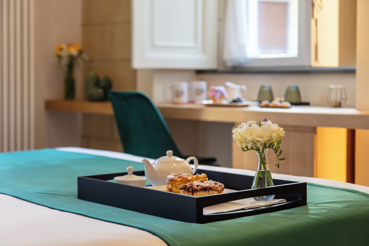 A neatly arranged breakfast tray with a teapot, a pastry, and a vase of flowers on a bed with a green bedspread.