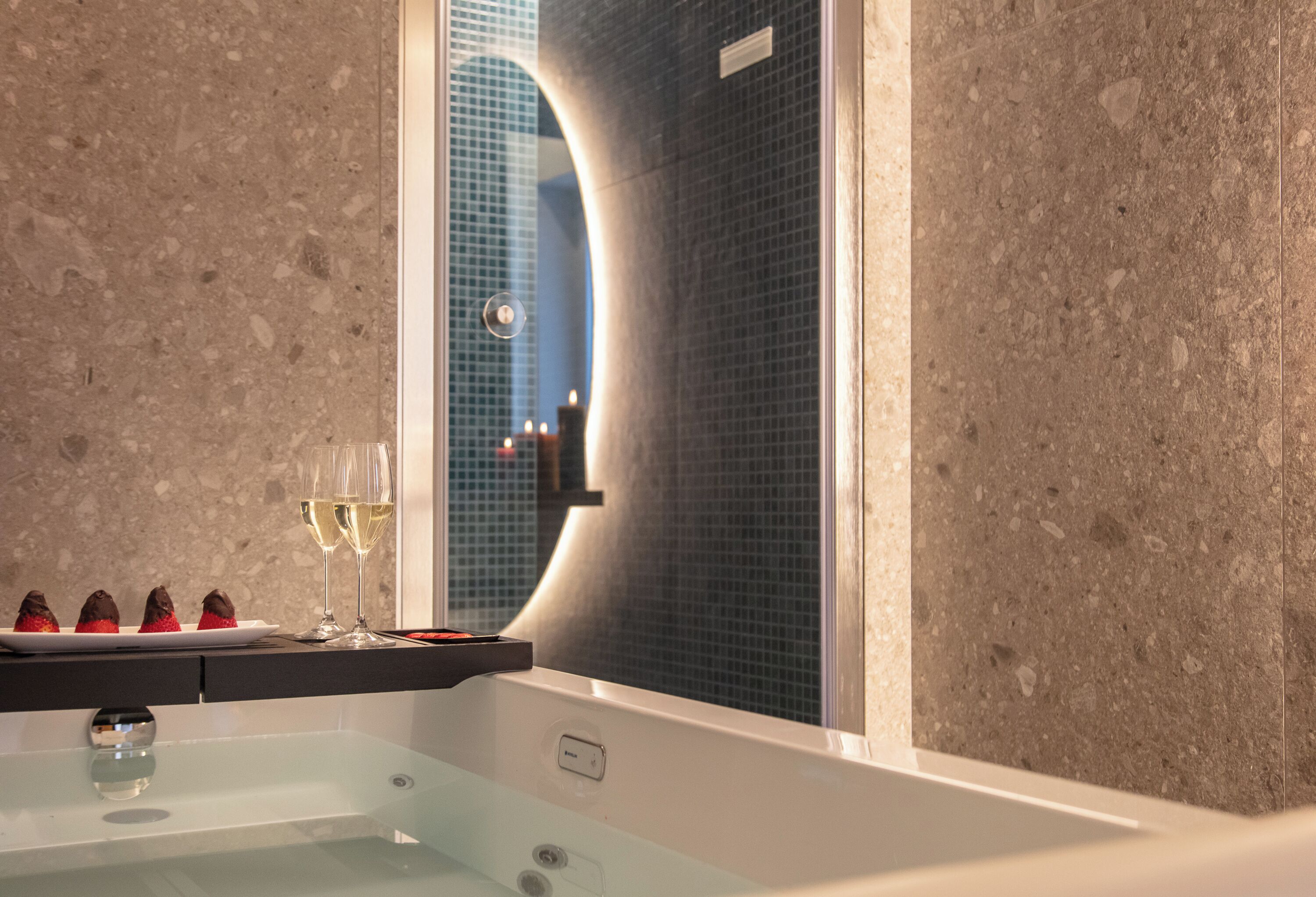 A modern bathroom with a large mirror, a glass bathtub, and a countertop with chocolate-covered strawberries and a glass of white wine.