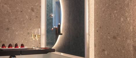A modern bathroom with a large mirror, a glass bathtub, and a countertop with chocolate-covered strawberries and a glass of white wine.