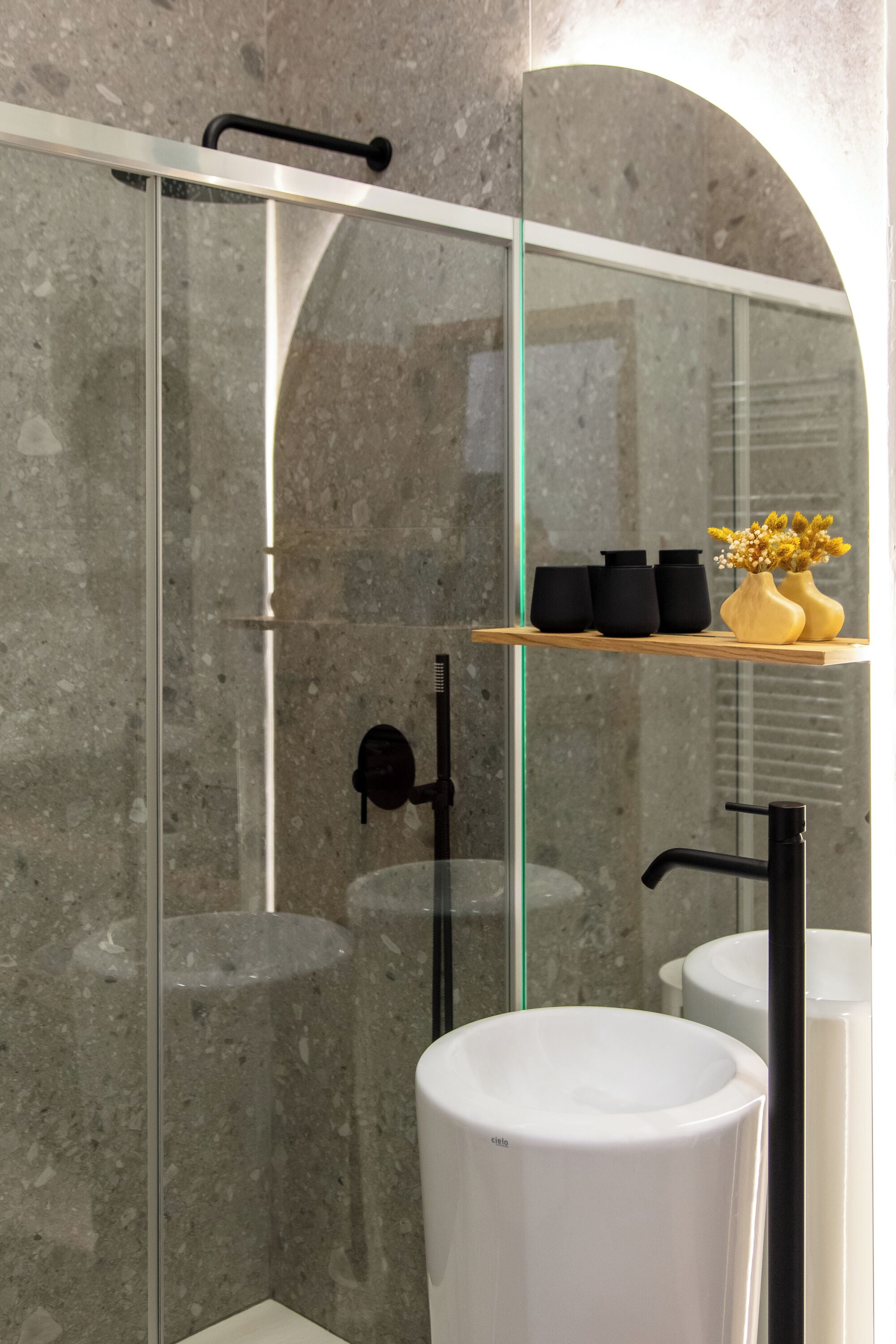Un baño moderno con ducha envuelta en vidrio, un estante de madera con jarrones negros y amarillos, y lavamanos blancos.