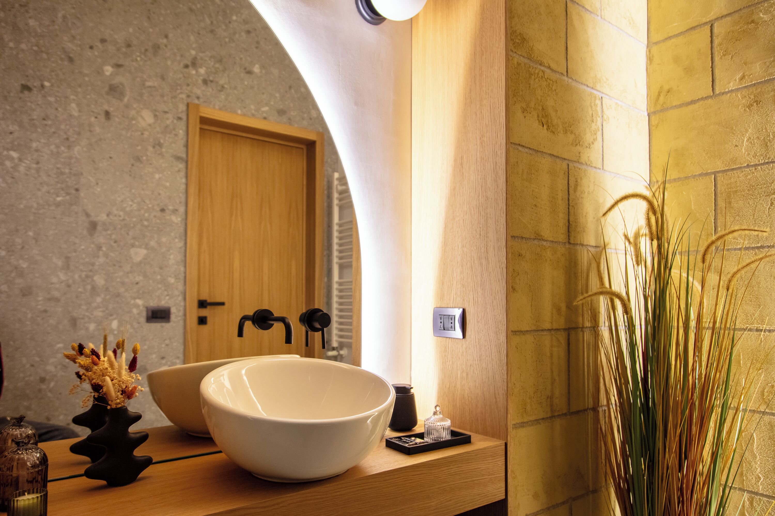 A bathroom with a large white sink, a wooden vanity, a mirror, and a vase with dried plants.
