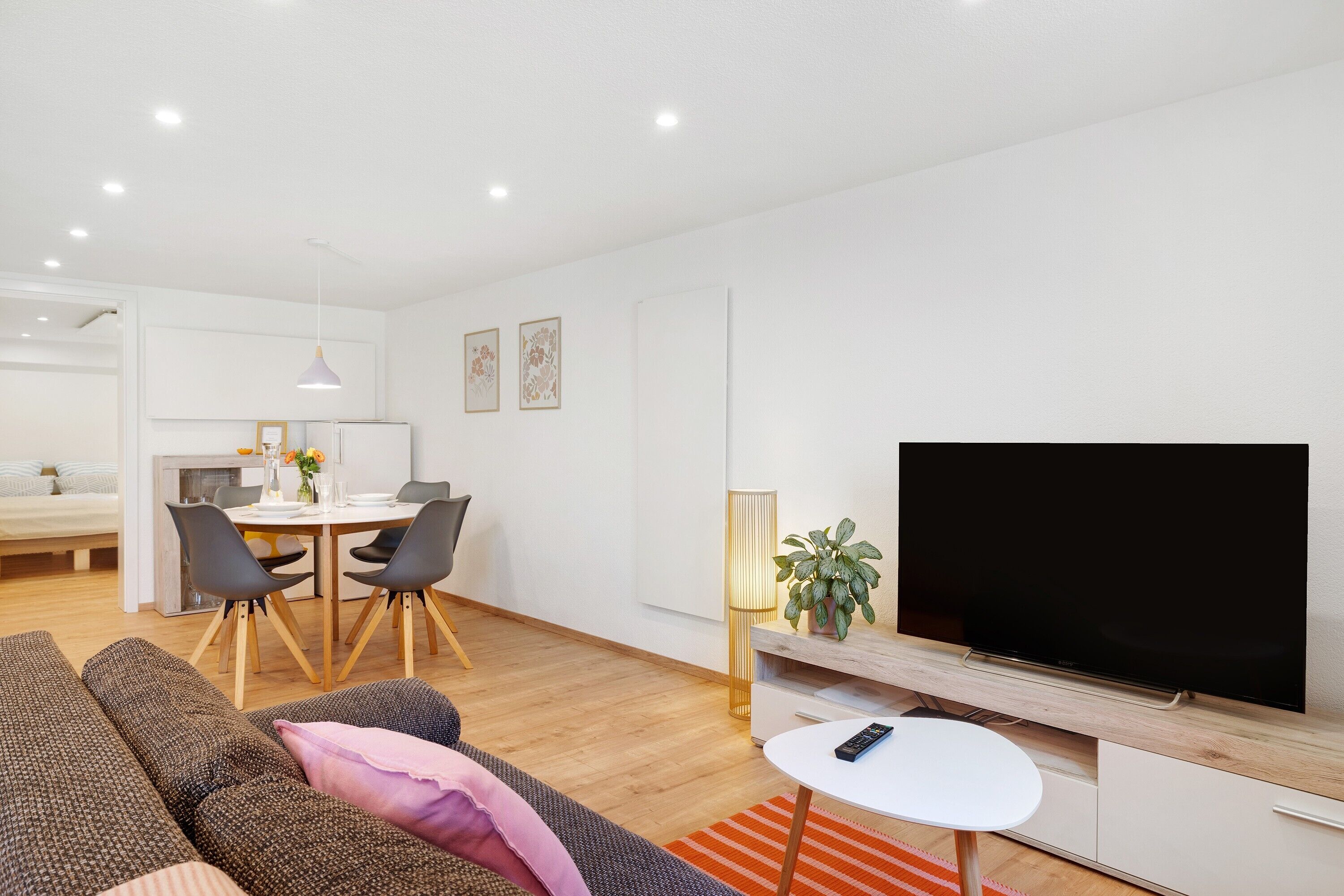 Ett modernt vardagsrum med en platt-TV, en matplats med bord och stolar samt en mysig sittgrupp med en soffa och ett litet vitt bord.