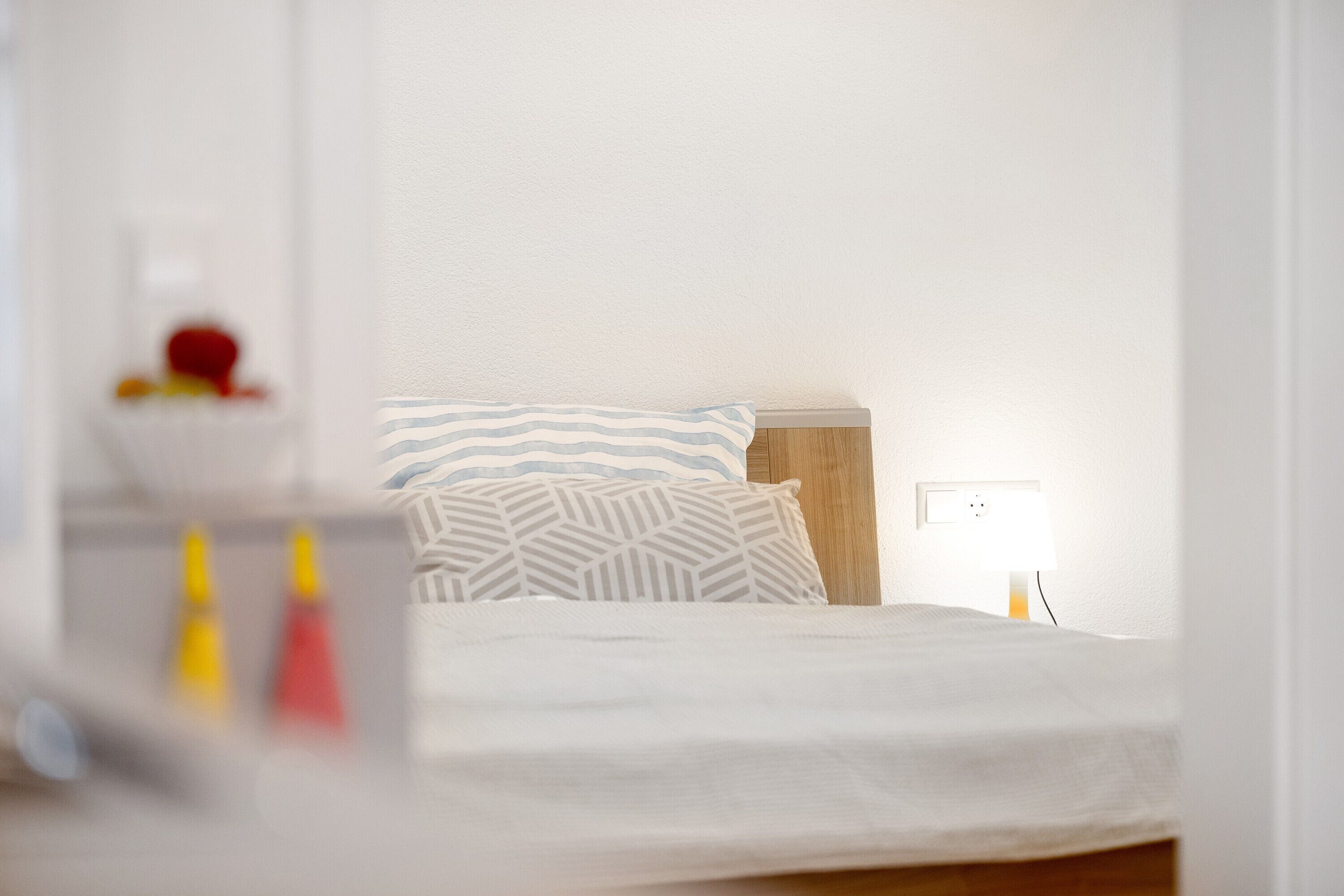 Una cama con un almohadón a rayas y un almohadón liso, una mesita de noche con una lámpara, y un enchufe de pared.