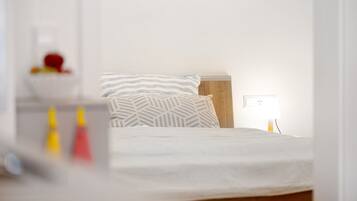 Una cama con un almohadón a rayas y un almohadón liso, una mesita de noche con una lámpara, y un enchufe de pared.
