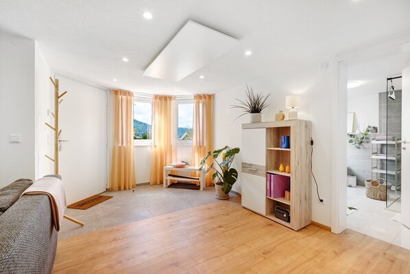 A modern living room with a sofa, a wooden coffee table, a small bench, a potted plant, a bookshelf, and a glass shower enclosure.