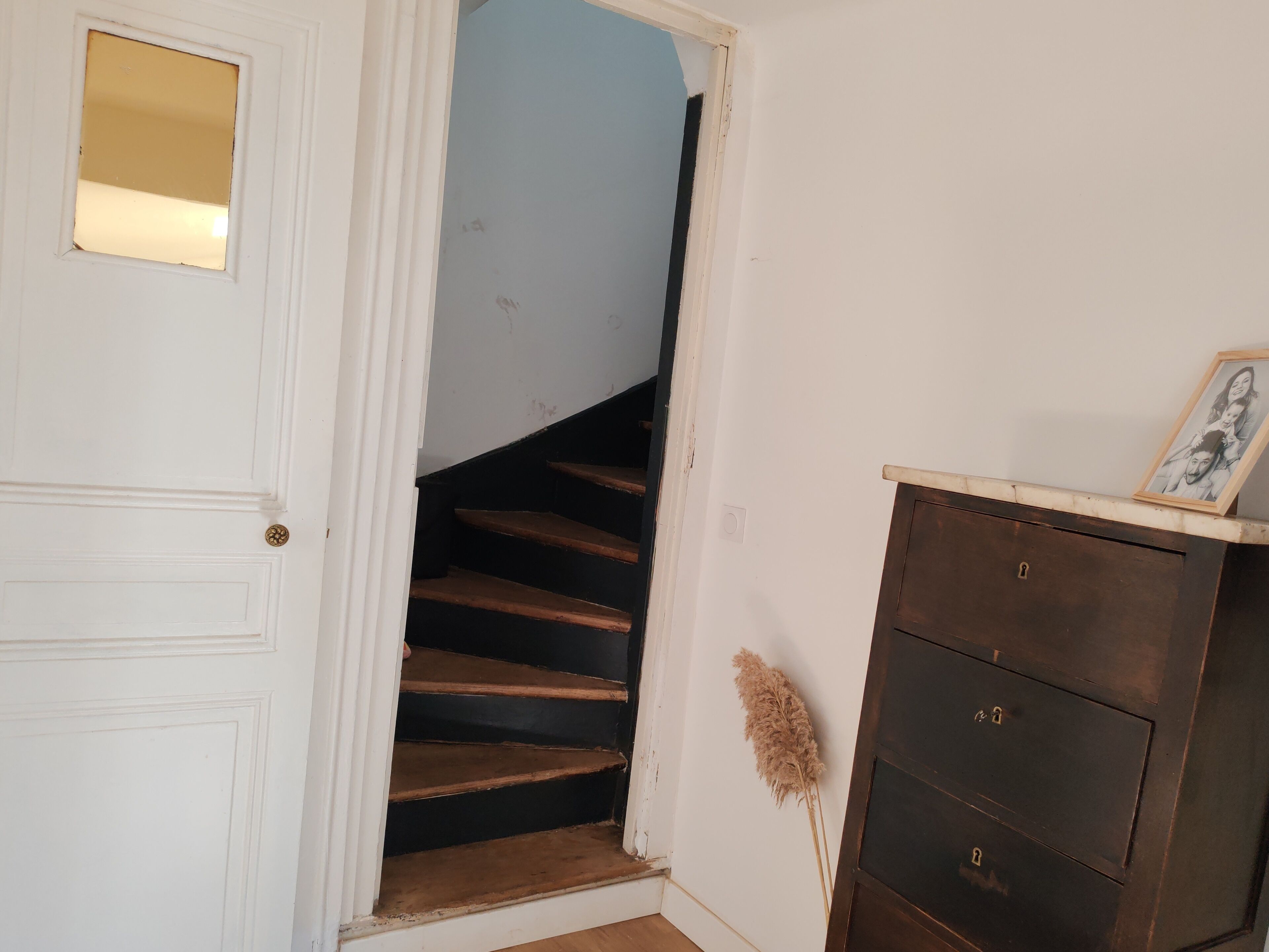 A white door with a small mirror, a wooden staircase, and a dark wooden dresser with a picture frame.