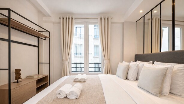 A hotel room with a bed, a wooden side table, a mirror, and a view of the outside. - 1628 Matignon (Paris)