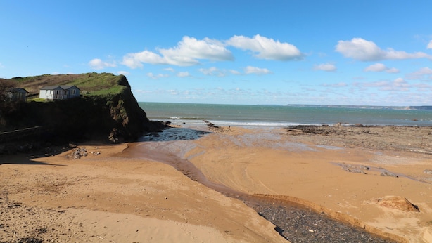Un paysage côtier avec une plage de sable, une falaise herbeuse et un petit bâtiment situé sur la falaise.