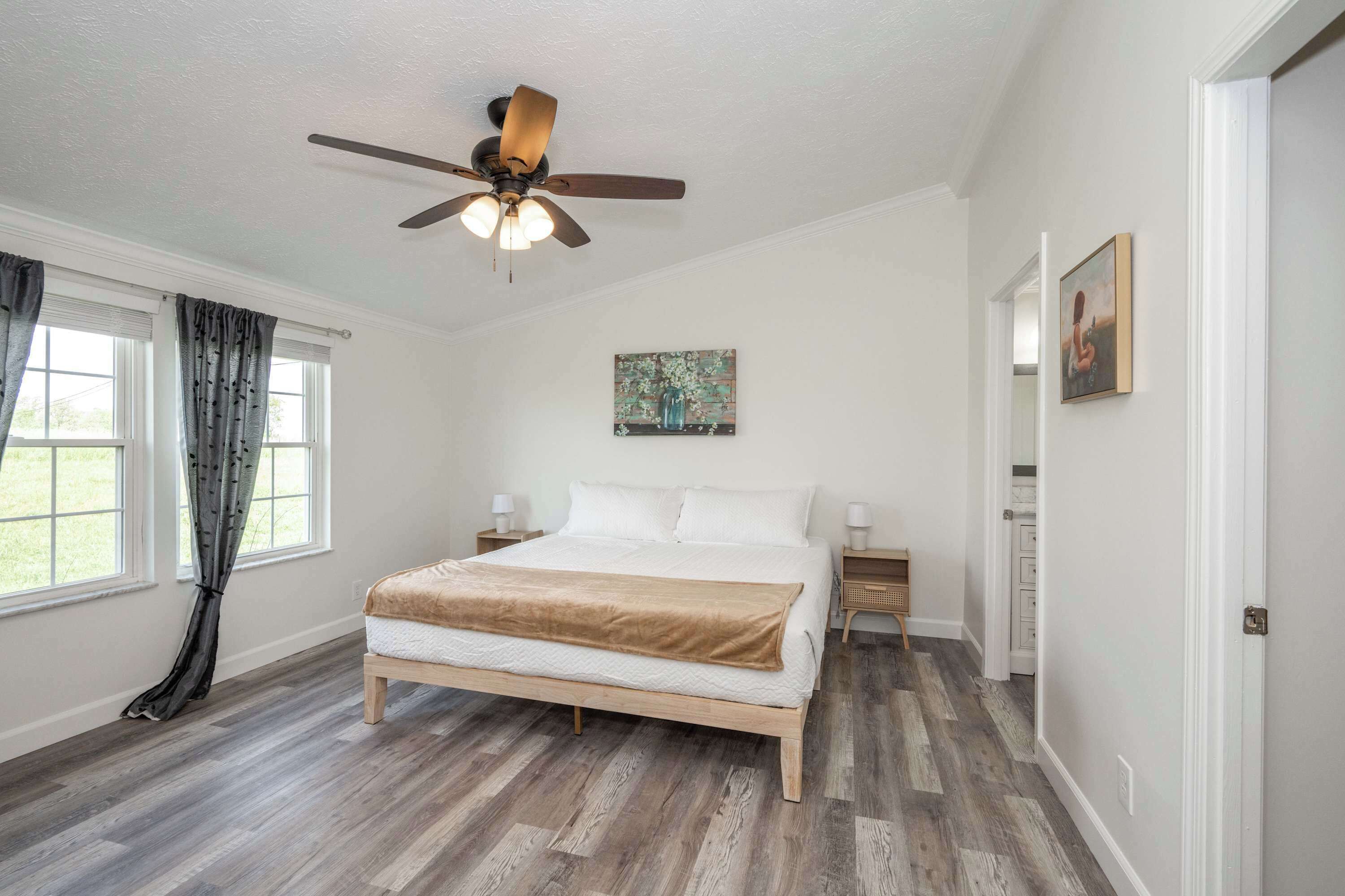 A bedroom with a wooden bed, a ceiling fan, a window with curtains, and a painting on the wall.