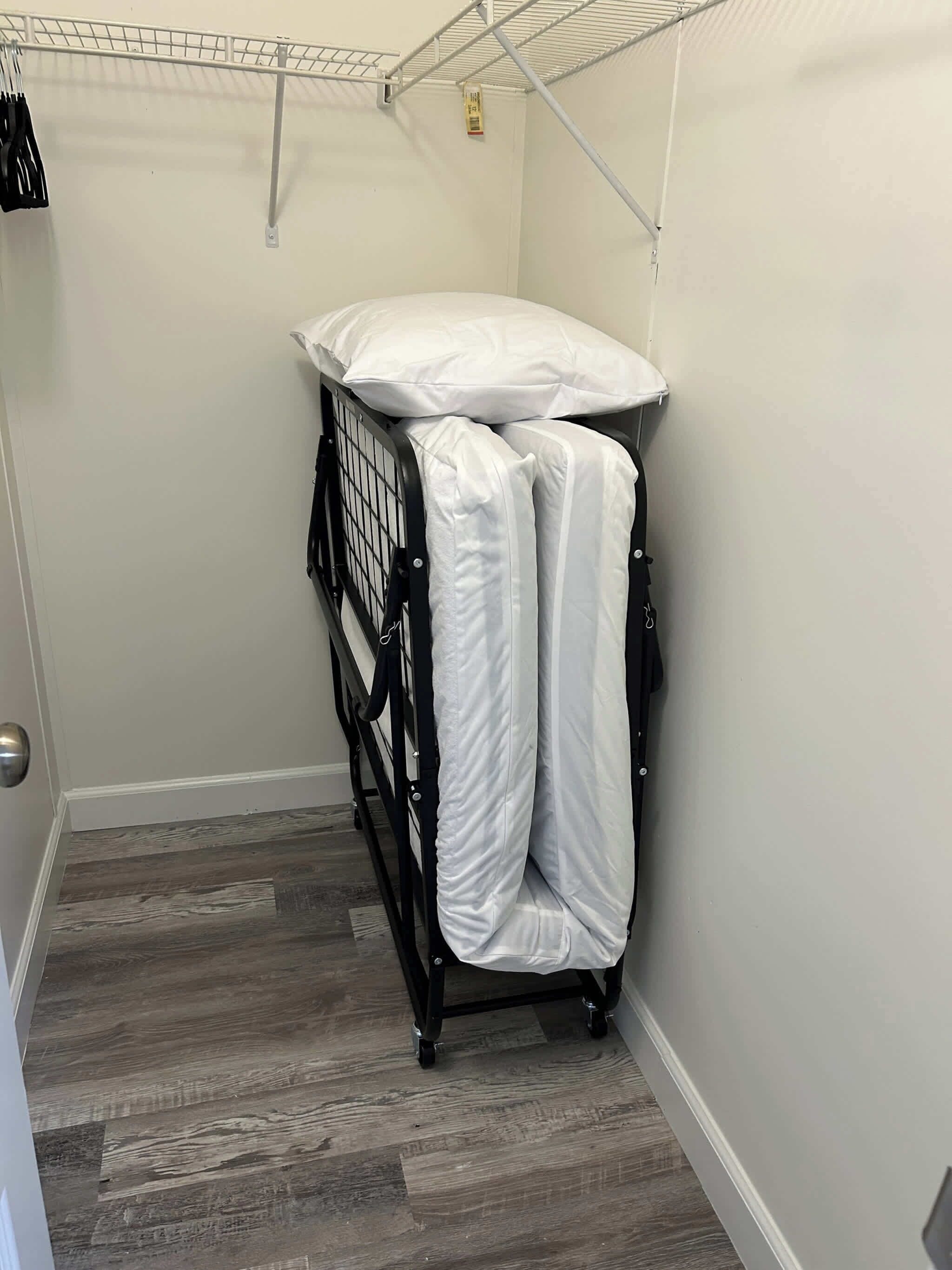 A compact room with a metal rack and a mattress on wheels, a door with a handle, and a wooden floor.