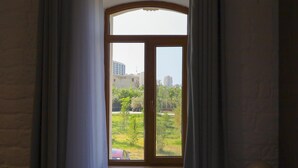 Una habitación con un gran ventanal arqueado, cortinas, una lámpara y vistas a edificios y zonas verdes al exterior.