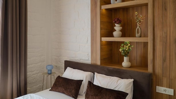 Un dormitorio con cama, dos almohadas, una mesita de noche, una lámpara y un estante de madera con objetos decorativos.