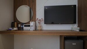 Una habitación de hotel moderna con un escritorio de madera, un televisor de pantalla plana, un espejo redondo, un refrigerador pequeño y una mesita de noche con un cojín blanco.
