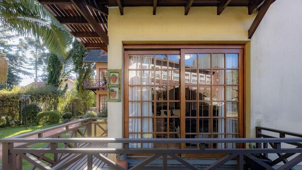 A wooden deck with a railing, a large window with multiple panes, and a balcony with potted plants. - Apartamento em Gramado próximo ao Parque Carrieri (Gramado)