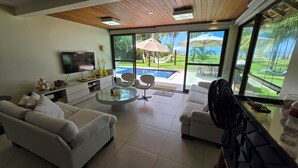 A modern living room with a TV, comfortable seating, a glass coffee table, and a view of a pool and outdoor area. - Bangalô Frente Mar no Carneiros Beach Resort bLP (Tamandaré)