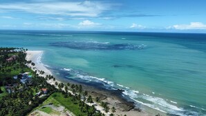 An aerial view of a coastal area with a beach, palm trees, and a small settlement with buildings. - Bangalô Frente Mar no Carneiros Beach Resort bLP (Tamandaré)